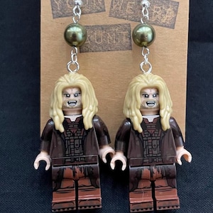 May include: A pair of unique earrings featuring Lego minifigure characters with long blonde hair and brown outfits. Each earring has a silver hook, a green bead, and a Lego figure. The earrings are displayed on a brown card with text.