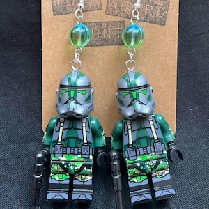 May include: A pair of handmade earrings featuring miniature green and gray soldier figures. The earrings have silver hooks, clear and green bead accents, and are attached to a brown card with the text "YOUR WEIRD HUNT".