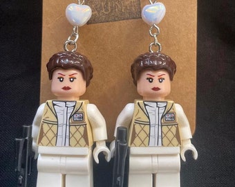 May include: A pair of dangle earrings featuring a Lego minifigure of Princess Leia in her white Rebel pilot outfit holding a blaster. Each earring has a silver heart charm and a silver hook.