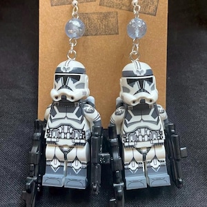 May include: Silver-tone drop earrings with white, grey, and black toy soldier figures and black rifles. Each earring has a light blue bead above the figure, suspended from a silver hook. The earrings are displayed on a brown card.