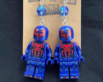 Puede incluir: Pendientes hechos a mano con figuras de Lego azules con detalles en rojo y negro. Los pendientes tienen ganchos plateados y cuentas azules iridiscentes. Se exhiben en una tarjeta marrón con el texto "YOUR WEIRD".