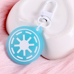 Puede incluir: Un llavero azul y blanco con el símbolo de la Alianza Rebelde de Star Wars. El llavero tiene un anillo de metal blanco y un clip de metal blanco.
