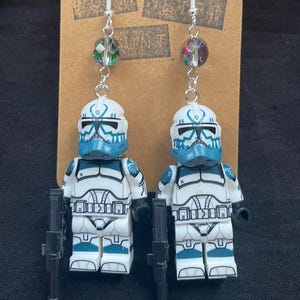 May include: Dangle earrings with white and blue toy soldier figures and black rifles. The earrings have silver hooks and colourful glass beads. They are displayed on a brown cardboard backing. The earrings are a fun, quirky accessory.