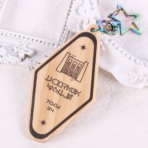 May include: Wooden keychain with a diamond-shaped tag featuring a black and white illustration of a building with a star above it. The tag also has the text "7000E 34" and a series of symbols. The keychain is attached to a rainbow-coloured star charm.