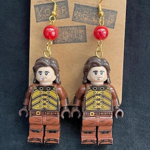 May include: A pair of handmade earrings featuring Lego minifigure charms. Each earring has a gold-colored hook, a red bead, and a Lego figure with brown hair and a brown and yellow outfit. The earrings are displayed on a brown card.