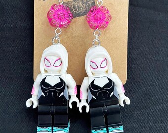 Puede incluir: Un par de pendientes con una minifigura Lego blanca de Spider-Gwen con un traje negro y detalles rosas. Los pendientes son plateados y tienen un dije de flor rosa en la parte superior.