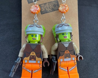 May include: A pair of earrings featuring Lego minifigures of a female character from Star Wars. The minifigures are wearing orange jumpsuits, brown vests, and grey helmets with goggles. The earrings are attached to silver hooks with orange flower beads.