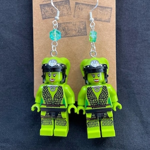 May include: A pair of green Lego minifigure earrings with black helmets and green and blue beads. The earrings are on a brown card with the text "Your Art".