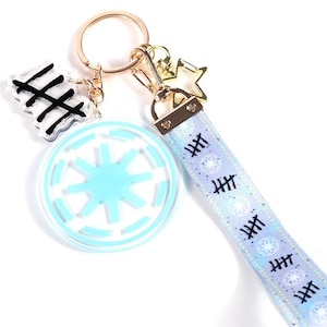 May include: A gold-coloured keyring with several charms. It includes a light blue and white Imperial symbol charm, a black and white charm with tally marks, and a light blue wrist strap with tally marks and star-like designs.