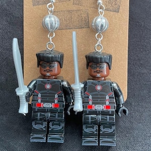 K&ouml;nnte beinhalten: Handgefertigte Ohrringe mit Lego-Minifiguren eines Charakters in einem schwarz-roten Outfit, der ein silbernes Schwert h&auml;lt. Die Ohrringe haben silberne Haken und sind an einer braunen Karte befestigt.