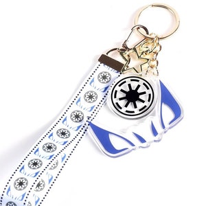 Pode incluir: Um chaveiro branco e azul com uma corrente dourada e um amuleto de estrela. O chaveiro apresenta um símbolo azul e branco da 501ª Legiao, uma unidade militar fictícia da franquia Star Wars.