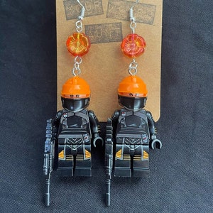 May include: A pair of earrings featuring Lego minifigures wearing black and orange armor, holding black guns, and a dangling orange flower charm.