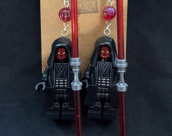 May include: A pair of dangle earrings featuring a Lego Darth Maul minifigure holding a red lightsaber. The earrings are silver with red beads and red lightsaber charms.