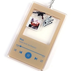 Può includere: Un portachiavi in acrilico trasparente a forma di lettore musicale con sfondo marrone. Il lettore ha il titolo della canzone "Run" di Taylor Swift ed Ed Sheeran. Il lettore contiene un'immagine di Han Solo di Star Wars e un'astronave Millennium Falcon.