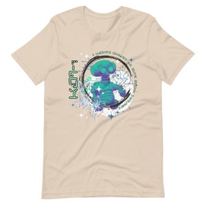 May include: A beige t-shirt with a graphic print of a blue and green robot-like figure in a circle with the text "I WOKE UP IN THE MATRIX" and "K-571-8" on the left side of the circle.