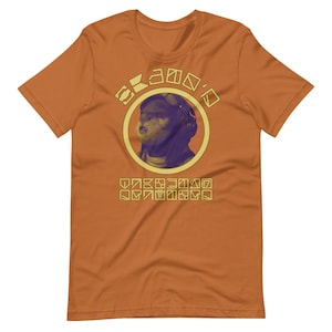 Op de afbeelding: Oranje T-shirt met korte mouwen met een gele en paarse illustratie van een harige creatuur met een helm en een bril. De tekst "KAMO'O" staat boven de illustratie en een reeks gele symbolen staan onder de illustratie.