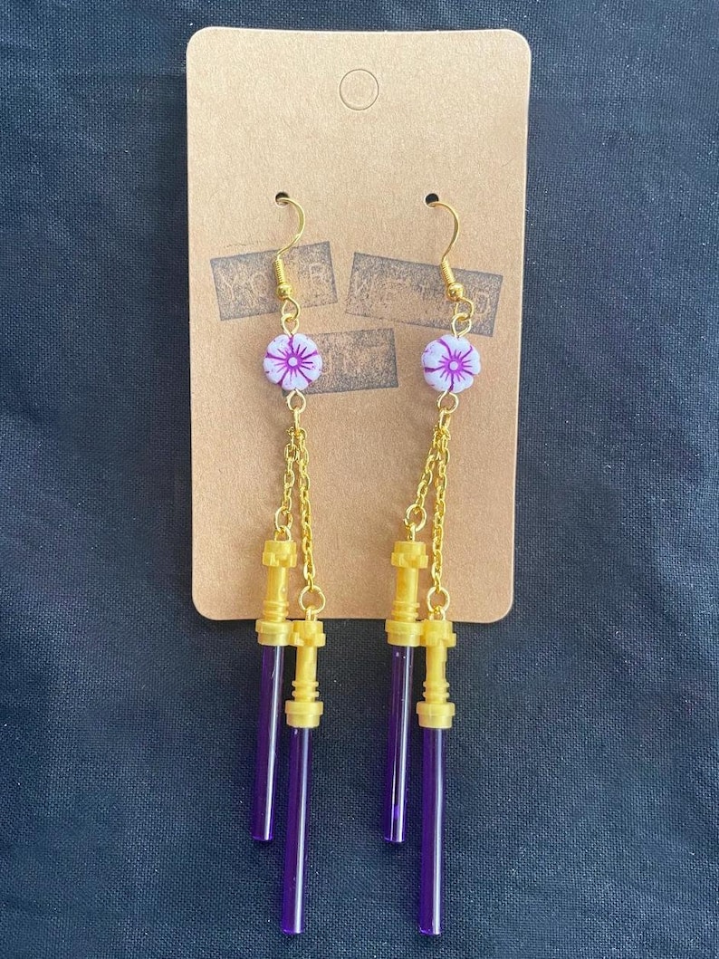 May include: A pair of dangle earrings with a purple and gold design. The earrings feature a small white flower charm with a pink center, followed by a gold chain and three purple lightsaber-shaped charms.