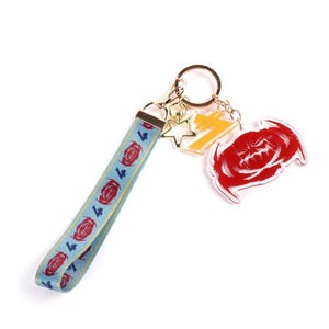 Può includere: Un portachiavi con un cinturino da polso azzurro con un motivo ripetuto di rose rosse e il numero 7. Il portachiavi ha un anello e ciondoli dorati, tra cui una stella, un fulmine giallo e una rosa rossa stilizzata.