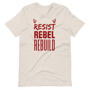 Könnte beinhalten: Cremefarbenes T-Shirt mit rotem Aufdruck "RESIST REBEL REBUILD" und zwei roten Teufelshörnern. Das Shirt hat einen Rundhalsausschnitt und kurze Ärmel.