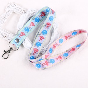 Puede incluir: Un cordón azul y rosa con un patrón repetido de símbolos rojos y azules. El cordón tiene un clip de metal plateado.