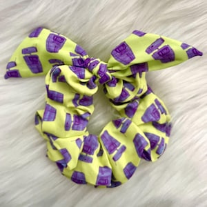 Op de afbeelding: Een gele scrunchie met een paars retro telefoonpatroon. De scrunchie is in een strik geknoopt.