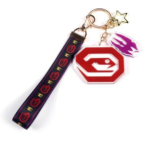 May include: A gold-toned keychain with a black and purple wrist strap featuring red and gold symbols. The keychain includes a red and white logo, a purple charm, and a gold star charm.