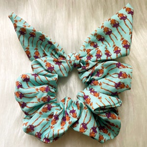 以下が含まれることがあります： 薄い青色のシュシュで、薄い青色の背景に茶色の図形が繰り返し描かれています。シュシュはリボン結びされています。