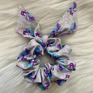 以下が含まれることがあります： リボンが付いた、紫と青の花柄のシュシュ。シュシュは柔らかな生地で作られており、ギャザーが寄せられています。