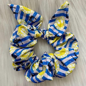 May include: A blue and white striped scrunchie with a yellow circular design and black silhouettes. The scrunchie has a bow-like top. The fabric is soft and gathered, creating a textured appearance. The accessory is designed for hair styling.
