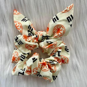 以下が含まれることがあります： クリーム色のシュシュ、オレンジと黒のシンボルが繰り返されるパターンが特徴です。 シュシュは布製で、ヘアスタイリング用にデザインされています。 リボンはアクセサリーに装飾的な要素を追加します。