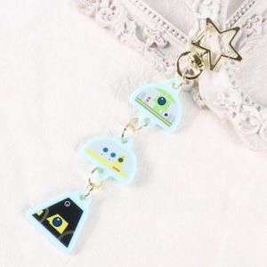 May include: A light blue keychain featuring three stylized robot-like charms. The charms are connected by small gold-colored chains and a gold star-shaped clasp. The charms have various geometric designs in white, yellow, and black.