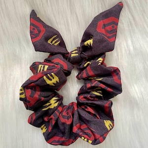 Puede incluir: Una scrunchie burdeos con lazo, con un patrón repetido de formas geométricas rojas y amarillas. Este accesorio para el cabello de tela está diseñado para atar el cabello. El lazo añade un toque decorativo.