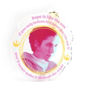 May include: A pink and gold round acrylic keychain with a quote from Star Wars: "Hope is like the sun. If you only believe in it when you can see it, you'll never make it through the night." The quote is surrounded by a gold circle and a picture of Princess Leia.