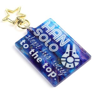 Può includere: Un portachiavi rettangolare con una chiusura dorata a forma di stella. Il portachiavi presenta un design marmorizzato blu e viola con testo bianco che recita "HAN SOLO, very this way to the top". È presente anche una grafica bianca del Millennium Falcon.