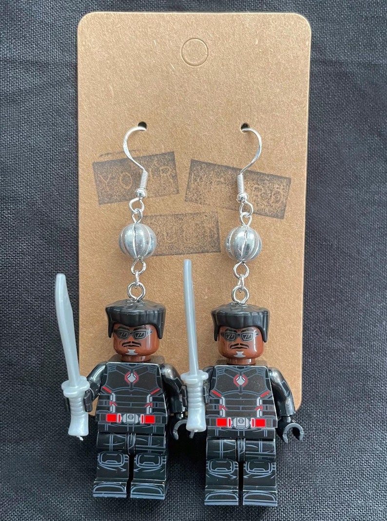 K&ouml;nnte beinhalten: Handgefertigte Ohrringe mit Lego-Minifiguren eines Charakters in einem schwarz-roten Outfit, der ein silbernes Schwert h&auml;lt. Die Ohrringe haben silberne Haken und sind an einer braunen Karte befestigt.