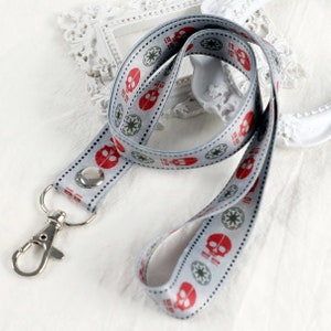 Pode incluir: Um cordão cinza e branco com um padrão repetido de caveiras vermelhas e símbolos de estrelas verdes. O cordão tem um clipe de metal prateado.