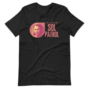 Op de afbeelding: Een zwart T-shirt met een roze cirkel met daarin een mannengezicht en de tekst "ask me about the SOL PATROL" in roze.
