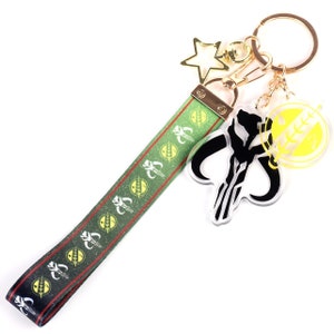 Op de afbeelding: Een groene en zwarte sleutelhanger met een bedel met het Mandalorian-symbool, een gouden sterbedel en een gouden ketting. De sleutelhanger heeft een groen lint met een herhalend patroon van het Mandalorian-symbool.