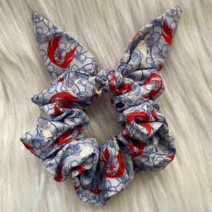 以下が含まれることがあります： 水色のシュシュ。花柄と赤いアクセントが特徴です。シュシュはリボンのようなデザインで、耳の形をした2つのエクステンションが付いています。生地には、小さな青い花と赤いシンボルの繰り返し模様があります。