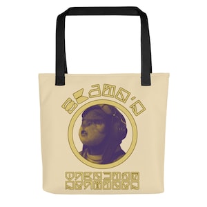 Op de afbeelding: Een beige tote bag met zwarte handvatten heeft een gele cirkel met het gezicht van een personage erin. De cirkel heeft de tekst "O-KAMO-O" eromheen. De tekst is in een gestileerd lettertype. De tas heeft ook een patroon van gele vierkanten met symbolen erin.