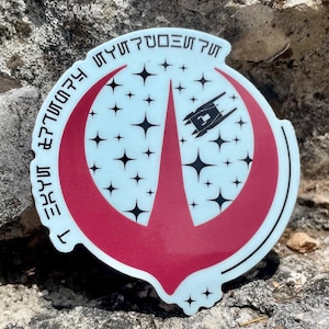 Può includere: Un adesivo rotondo con sfondo azzurro chiaro e un emblema rosso. L'emblema presenta un design stilizzato con una nave e stelle. L'adesivo ha testo attorno al bordo in un carattere unico.