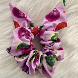 Op de afbeelding: Een paarse scrunchie met een herhalend patroon van roze, groene en paarse vruchten. De scrunchie is gemaakt van een zachte, rekbare stof.