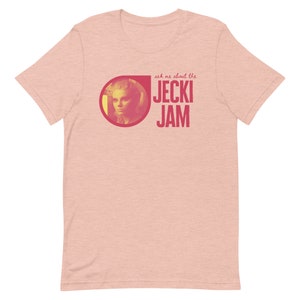 Op de afbeelding: Een roze heather t-shirt met een rode cirkelvormige afbeelding met een vrouwengezicht en de tekst "ask me about the JECKI JAM".