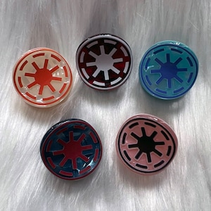 Op de afbeelding: Vijf ronde, plastic haarclips met een wit sterburst-ontwerp in een zwarte cirkel. De sterburst is in verschillende kleuren: rood, wit, blauw, roze en donkerrood.