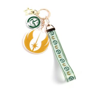 Peut inclure: Un porte-clés doré avec plusieurs breloques. Les breloques comprennent une étoile dorée, un emblème circulaire vert et blanc et un emblème jaune et blanc. Une lanière verte et blanche avec un motif répétitif est attachée.