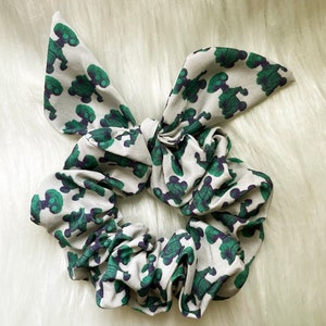 以下が含まれることがあります： 緑と黒の宇宙人の模様がついた白いシュシュ。シュシュはリボン結びされています。