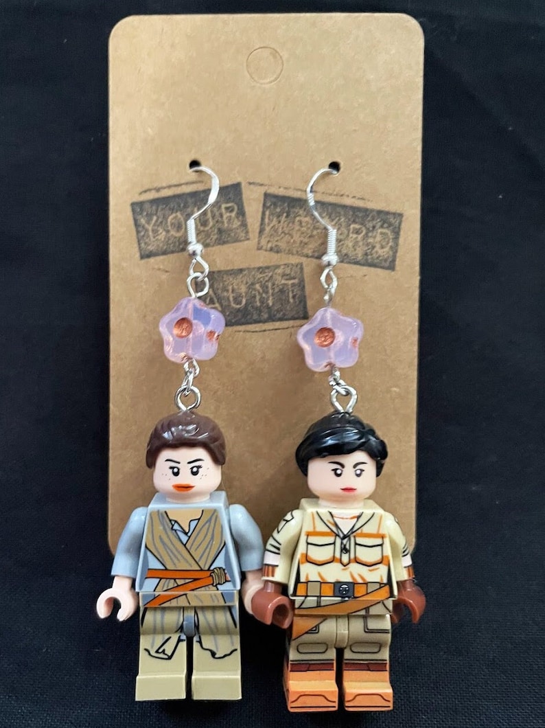 K&ouml;nnte beinhalten: Ein Paar Ohrringe mit Lego-Minifiguren von Rey und einer weiblichen Figur aus Star Wars. Die Minifiguren sind an silbernen Ketten mit rosa bl&uuml;tenf&ouml;rmigen Perlen befestigt.