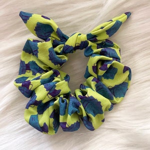 以下が含まれることがあります： 緑と青のシュシュで、漫画のようなキャラクターが繰り返し描かれたパターンがあります。シュシュはリボン結びされています。