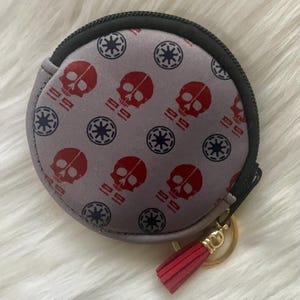 Op de afbeelding: Een ronde, grijze portemonnee met een zwarte ritssluiting en een rode kwast. De portemonnee is bedrukt met rode schedels en blauwe sterrenpatronen. Een goudkleurige sleutelhanger is bevestigd.