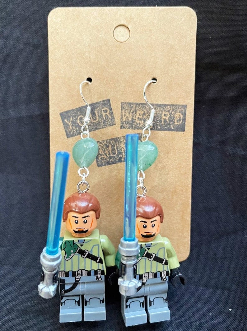 Pu&ograve; includere: Un paio di orecchini con minifigure Lego che tengono spade laser blu chiaro. Ogni minifigure indossa un abito verde e grigio e ha i capelli castani. Gli orecchini sono decorati con perline verdi a forma di cuore.